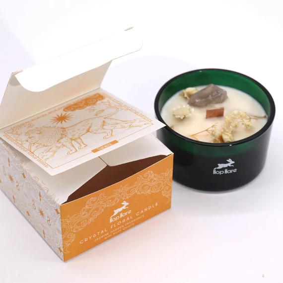 Az Oroszlán kristálygyertya hegyikristállyal és szárított virágokkal zöld üveg tégelyben, mellette a Hop Hare narancssárga tarot-stílusú díszdoboza.